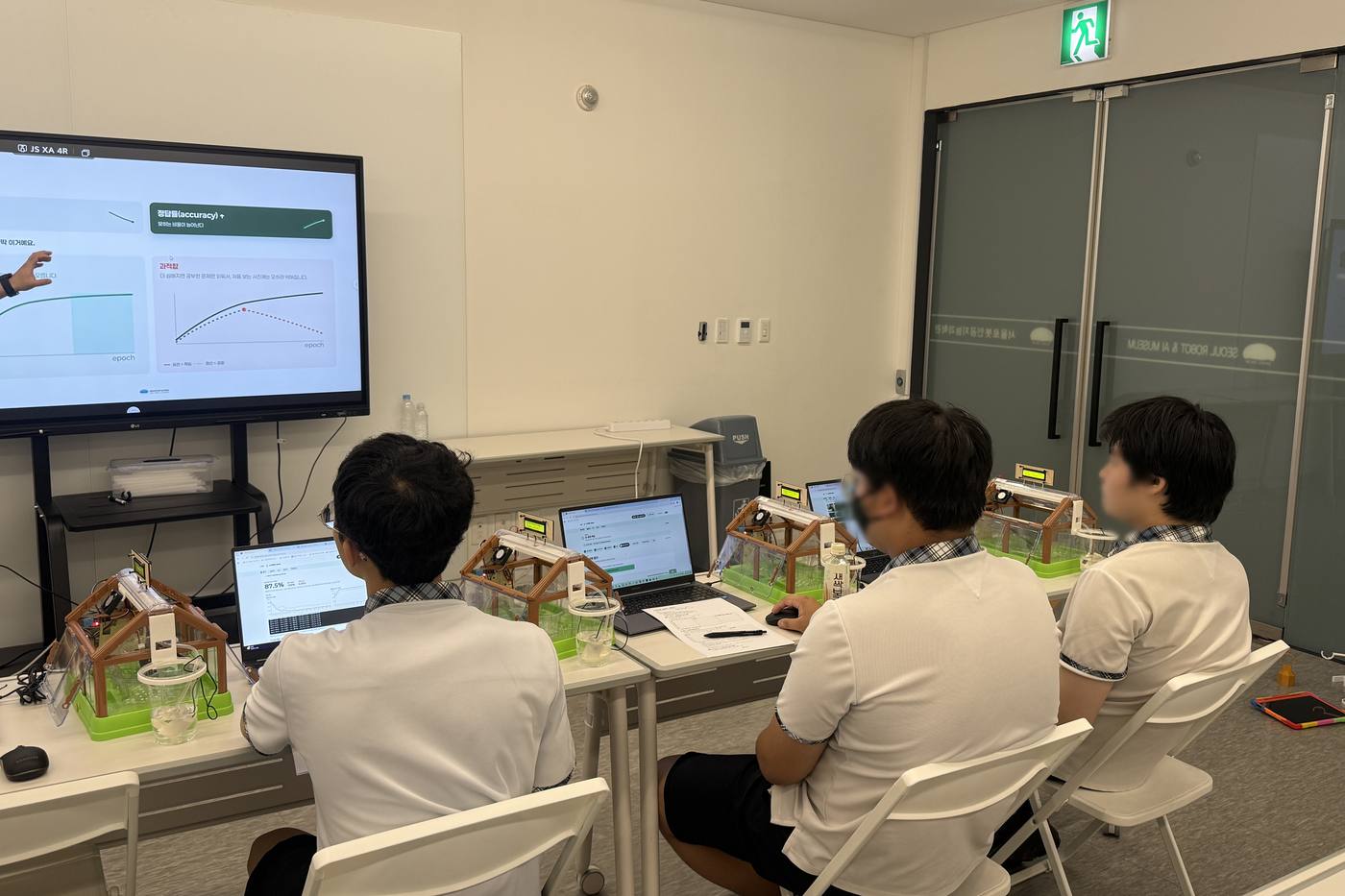 Demo class in August 2026 on the museum's education floor: students watch their own in-browser model training reach 87.5% accuracy, each with a mini smart-farm sensor rig beside them.